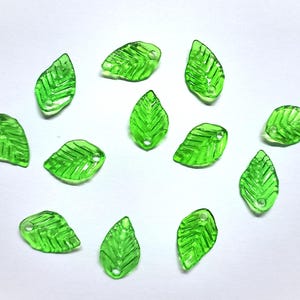 Op de afbeelding: Een verzameling doorschijnende groene blad-vormige kralen. Elke kraal heeft een klein gaatje om te rijgen en gedetailleerde nerven. De kralen zijn verspreid op een witte ondergrond, wat een natuurlijke, organische uitstraling creëert. Ideaal voor het maken van sieraden en knutselprojecten.