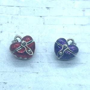 Op de afbeelding: Twee hartvormige hangerbedels, één rood en één paars, met een zilveren sluiting en een zilveren lus om aan een ketting of armband te bevestigen.