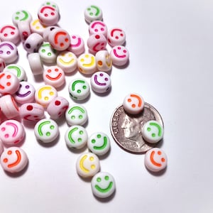 May include: A collection of small, round beads with smiley faces in various colors, including pink, green, orange, and purple. The beads are scattered on a white surface, with a US dime for size comparison. These beads are suitable for jewelry making and crafts.