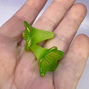 May include: Two translucent green flower-shaped beads with gold accents. The beads are held in a person's hand. The beads have a bell shape with detailed petal edges. The gold accents are on the edges of the petals.