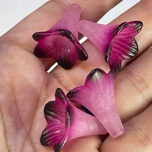 May include: Four pink and purple flower-shaped glass beads with black accents. The beads are translucent and have a frosted finish.