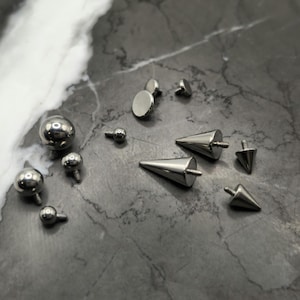 May include: A collection of silver-toned body jewelry, including balls, spikes, and flat studs. The jewelry is arranged on a gray marble surface.