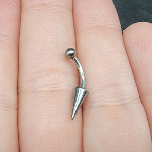 May include: A silver-colored belly button ring with a curved barbell design. The top features a small ball, and the bottom has a cone-shaped spike. The jewelry is shown against a neutral background.