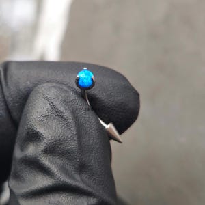 May include: A blue opal gemstone set in a silver barbell piercing with a pointed end.