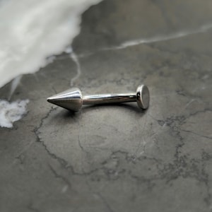 May include: A silver barbell with a pointed end and a flat circular end. The barbell is lying on a gray surface.