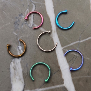 May include: Six nose rings in different colors, including pink, blue, silver, gold, green, and purple. The rings are all made of metal and have a small bead at the end.