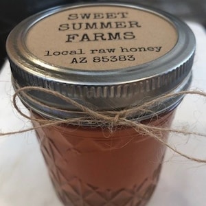 May include: A glass jar with a brown label that reads "Sweet Summer Farms, local raw honey, AZ 85383". The jar is tied with twine and has a textured surface.
