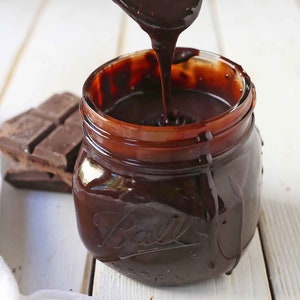 Puede incluir: Un tarro de vidrio lleno de salsa de chocolate espesa y oscura. La salsa se vierte de una cuchara. Una barra de chocolate es visible en el fondo.