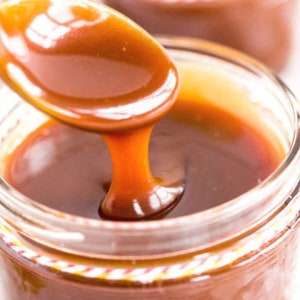 Puede incluir: Primer plano de un tarro lleno de salsa de caramelo. Una cuchara de caramelo se sostiene sobre el tarro, con la salsa espesa y brillante goteando de vuelta al recipiente. El caramelo es de un rico color marrón dorado.