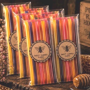 May include: Packets of honey sticks in various colors, including yellow, orange, pink, and purple, are displayed. Each packet has a circular label with a bee illustration and the text "Sweet Summer Farms." A wooden box with "Pure Raw Honey" is in the background.