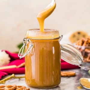 Puede incluir: Un tarro de vidrio lleno de salsa de caramelo marrón dorado. La salsa se vierte de una cuchara en el tarro. El tarro tiene un cierre de metal y una tapa de vidrio.