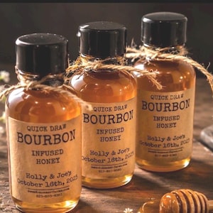 Puede incluir: Tres pequeñas botellas de vidrio llenas de líquido color ámbar, cada una con una tapa negra y una etiqueta que dice "Quick Draw Bourbon Infused Honey". Las etiquetas también incluyen "Holly & Joe's October 16th, 2025."