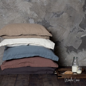Può includere: Una pila di cinque federe in lino in diverse tonalità di marrone, blu e bianco. Le federe sono disposte su una superficie di legno davanti a un muro di cemento grigio. Un piccolo vassoio con una bottiglia di vetro di latte e un bicchiere di latte si trova a destra dell'immagine. Il testo "Wonder Linen" è stampato sul vassoio.
