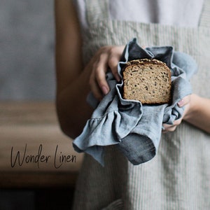 May include: A woman wearing a light blue and white striped linen apron holds a slice of bread wrapped in a blue linen tea towel. The text "Wonder Linen" is visible in the lower left corner of the image.