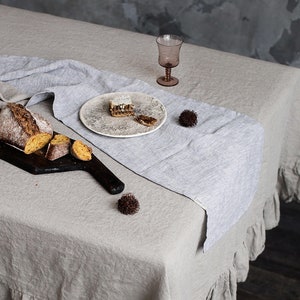 May include: A light grey linen tablecloth with a ruffled edge, a grey linen runner, a wooden chopping board with sliced bread, a plate with a piece of cake, a glass of wine, and two small decorative pinecones. The text "Wonder Linen" is visible in the bottom left corner of the image.