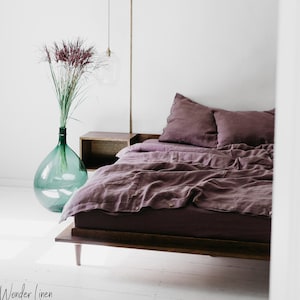 May include: A bedroom scene featuring a bed with plum-colored linen bedding and pillows. A large, clear glass vase with tall, dried plants sits beside the bed. The room has white walls and a wooden nightstand. The text "Wonder Linen" is visible.