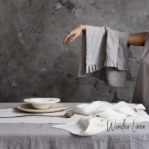 May include: A gray linen tablecloth with a white border. The tablecloth is set with a white ceramic bowl and a white linen napkin. The text "Wonder Linen" is visible in the lower right corner of the image.