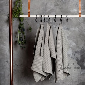 K&ouml;nnte beinhalten: Drei gestreifte Leinen-Geschirrt&uuml;cher h&auml;ngen an Haken, die an einer wei&szlig;en Metallstange befestigt sind. Die T&uuml;cher h&auml;ngen an einer grauen Betonwand. Der Text "Wonder Linen" ist in der rechten unteren Ecke des Bildes sichtbar.