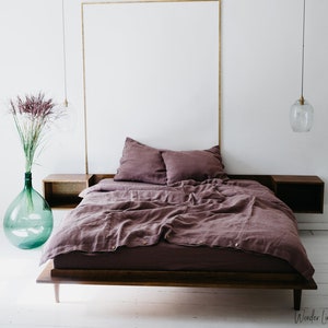 May include: A bedroom scene featuring a low-profile bed with a dark wood frame and dusty rose bedding. Two matching pillows rest on the bed. A large, clear glass vase with tall, dried flowers sits to the left. Two pendant lights hang from the ceiling.
