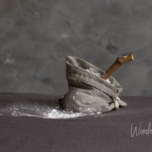 Op de afbeelding: Een grijze en witte gestreepte linnen tas met een houten stok erin. De tas ligt op een grijs oppervlak en er is wit poeder omheen. De tekst "Wonder Linen" is zichtbaar in de rechterbenedenhoek van de afbeelding.