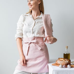 May include: A woman wearing a white shirt and a pink linen apron stands in front of a white wall. She is holding a pink linen towel in her right hand. A table with a white tablecloth is in the background, with a loaf of bread, garlic, and a bottle of olive oil on it.