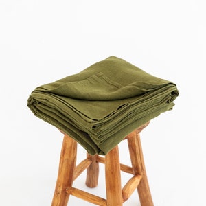 Könnte beinhalten: Ein Stapel gefalteter olivgrüner Leinenstoff auf einem Holzhocker. Der Stoff ist ordentlich gefaltet und hat eine weiche, natürliche Textur. Wonder Linen steht in weißer Schrift in der unteren rechten Ecke des Bildes.