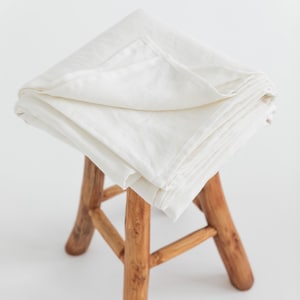 Pode incluir: Um tecido de linho branco dobrado e drapeado sobre um banquinho de madeira. O tecido é liso e tem uma textura natural. O banquinho é de madeira castanho claro e tem um design simples.