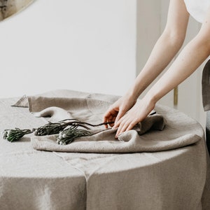 Op de afbeelding: Een persoon dekt een tafel met een linnen tafelkleed en een gestreepte linnen schort. Het tafelkleed is beige en het schort is grijs met witte strepen. De tekst "Wonder Linen" is zichtbaar in de linkerbenedenhoek van de afbeelding.