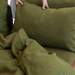  May include: Olive green linen bedding set, including a pillow and a duvet cover. The fabric has a soft, textured appearance. The bedding is displayed on a bed, with a person's hands adjusting a pillow. The Wonder Linen logo is visible.