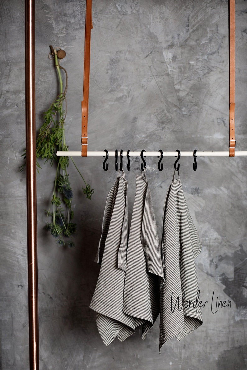 K&ouml;nnte beinhalten: Drei gestreifte Leinen-Geschirrt&uuml;cher h&auml;ngen an Haken, die an einer wei&szlig;en Metallstange befestigt sind. Die T&uuml;cher h&auml;ngen an einer grauen Betonwand. Der Text "Wonder Linen" ist in der rechten unteren Ecke des Bildes sichtbar.