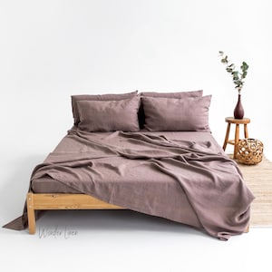 Può includere: Un set di lenzuola in lino malva su una struttura letto in legno. Il lenzuolo è ripiegato all'indietro per mostrare il lenzuolo con angoli elastici sottostante. Il letto è in una stanza bianca con un piccolo sgabello di legno e un vaso di verde in background.