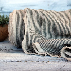K&ouml;nnte beinhalten: Eine Rolle aus nat&uuml;rlichem Leinenstoff mit schwarzen Streifen. Der Stoff hat einen rohen, unfertigen Rand. Der Stoff ist gefaltet und auf einer Holzoberfl&auml;che pr&auml;sentiert. Der Text "Wonder Linen" ist im Bild sichtbar.
