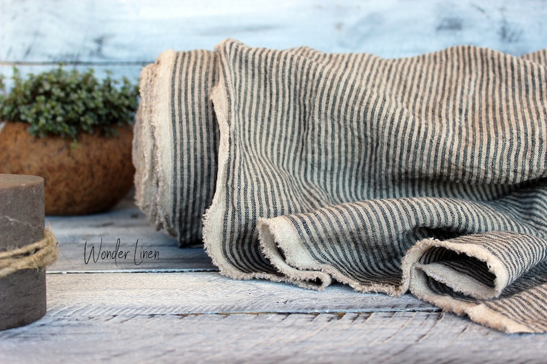 K&ouml;nnte beinhalten: Eine Rolle aus nat&uuml;rlichem Leinenstoff mit schwarzen Streifen. Der Stoff hat einen rohen, unfertigen Rand. Der Stoff ist gefaltet und auf einer Holzoberfl&auml;che pr&auml;sentiert. Der Text "Wonder Linen" ist im Bild sichtbar.