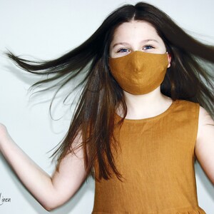 May include: A young person wearing a mustard yellow linen dress and a matching face mask. The person's hair is long and brown and is flowing in the wind. The background is a plain white wall. "Wonder Linen" is written in black text at the bottom of the image.