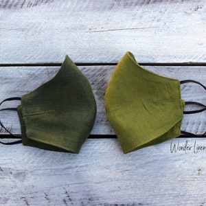 May include: Two green linen face masks with black elastic ear loops. The masks are laid out on a white wooden surface. The text "Wonder Linen" is visible in the bottom right corner of the image.