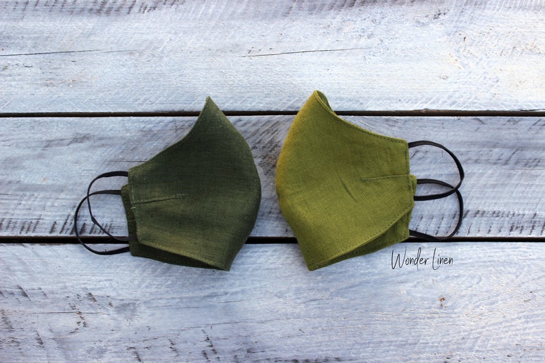 May include: Two green linen face masks with black elastic ear loops. The masks are laid out on a white wooden surface. The text "Wonder Linen" is visible in the bottom right corner of the image.
