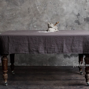 May include: A dark grey linen tablecloth draped over a wooden table with wheels. The tablecloth is textured and has a slight sheen. The table is in a room with a grey concrete wall.