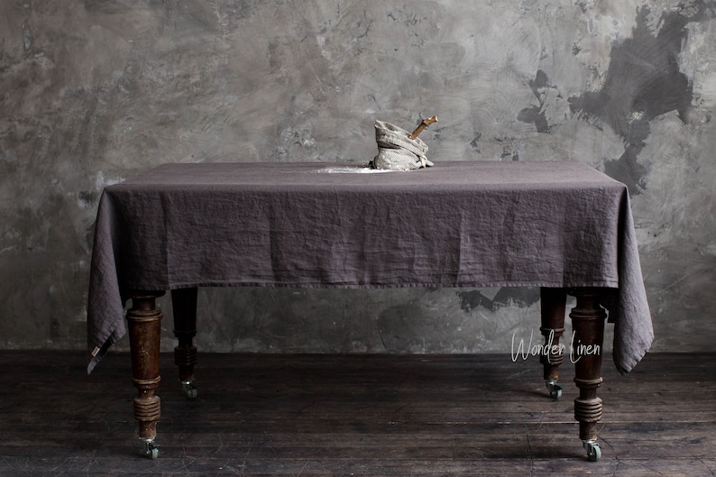 May include: A dark grey linen tablecloth draped over a wooden table with wheels. The tablecloth is textured and has a slight sheen. The table is in a room with a grey concrete wall.