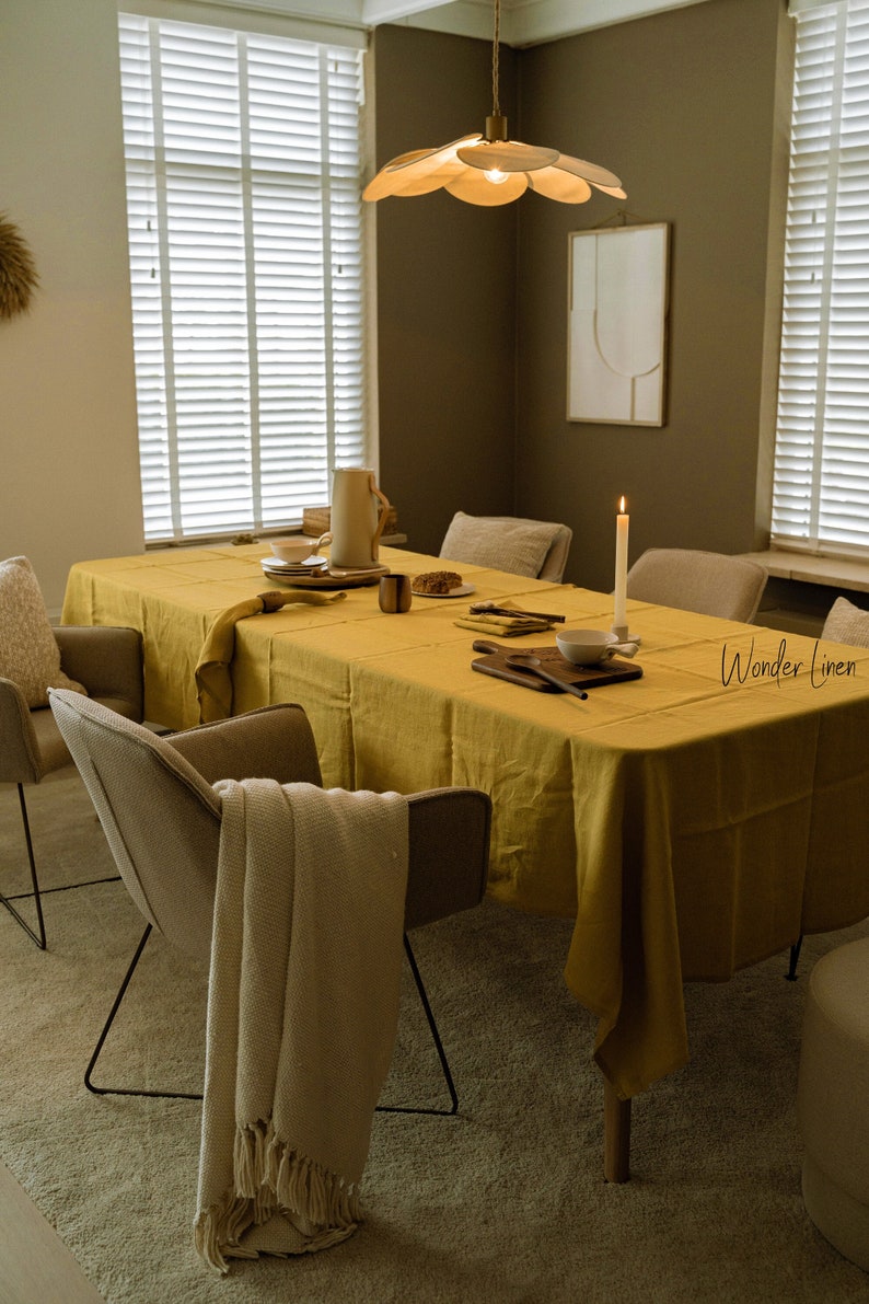 May include: A yellow linen tablecloth covers a wooden dining table with six chairs. The table is set with dishes, silverware, and a wooden cutting board. A white blanket is draped over one of the chairs. The room has a brown wall with a white framed print and a wooden ceiling fan with a light fixture.