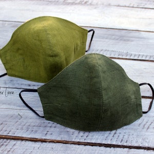 May include: Two green linen face masks with black elastic ear loops. The masks are on a white wooden surface. The text "Wonder Linen" is visible in the lower left corner of the image.