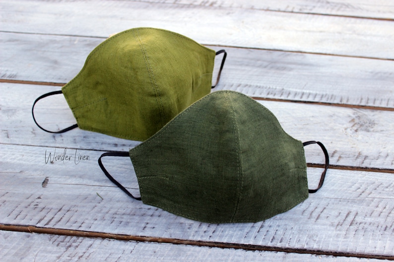 May include: Two green linen face masks with black elastic ear loops. The masks are on a white wooden surface. The text "Wonder Linen" is visible in the lower left corner of the image.