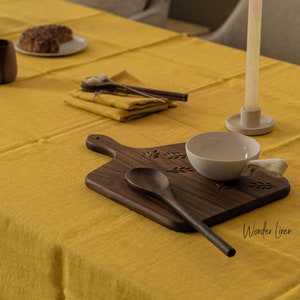 May include: A yellow linen tablecloth with a wooden cutting board, a small white bowl, a wooden spoon, and a lit candle on a white candle holder. The text "Wonder Linen" is visible in the bottom right corner of the image.