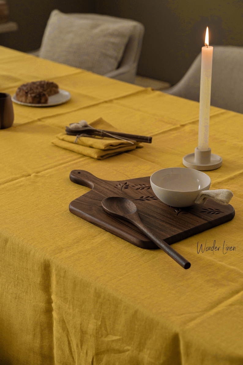 May include: A yellow linen tablecloth with a wooden cutting board, a small white bowl, a wooden spoon, and a lit candle on a white candle holder. The text "Wonder Linen" is visible in the bottom right corner of the image.