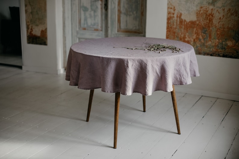 May include: A round table with a light purple linen tablecloth and wooden legs. The table is set on a white wooden floor.