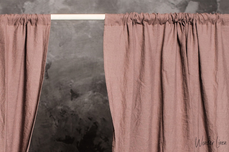 Puede incluir: Un par de cortinas de lino rosa polvoriento colgando de una barra blanca contra una pared gris texturizada. Las cortinas est&aacute;n reunidas en la parte superior y tienen un aspecto relajado y casual. El texto "Wonder Linen" es visible en la esquina inferior derecha de la imagen.