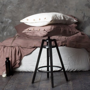 May include: A bed with a mauve linen duvet cover and pillows, accented with wooden buttons. A black metal stool is in the foreground, and a dark glass bottle and small container sit on the wooden floor.