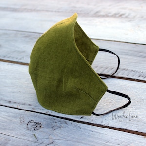 May include: A green linen face mask with black elastic ear loops. The mask is folded and lying on a white wooden surface. The text "Wonder Linen" is visible in the lower right corner of the image.