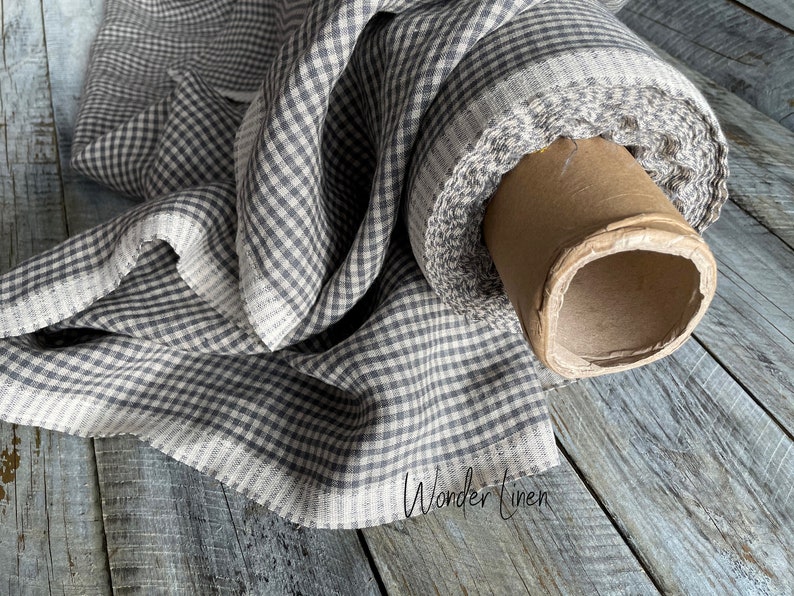 May include: A roll of gray and white gingham linen fabric. The fabric is folded and rolled on a cardboard tube. The fabric is on a wooden surface.