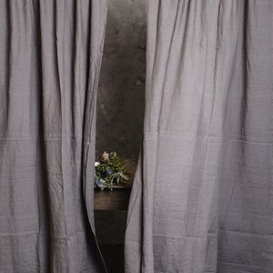 Puede incluir: Dos cortinas de lino gris colgadas de una barra blanca. Las cortinas est&aacute;n ligeramente corridas para revelar una mesa de madera con un ramo de flores secas.