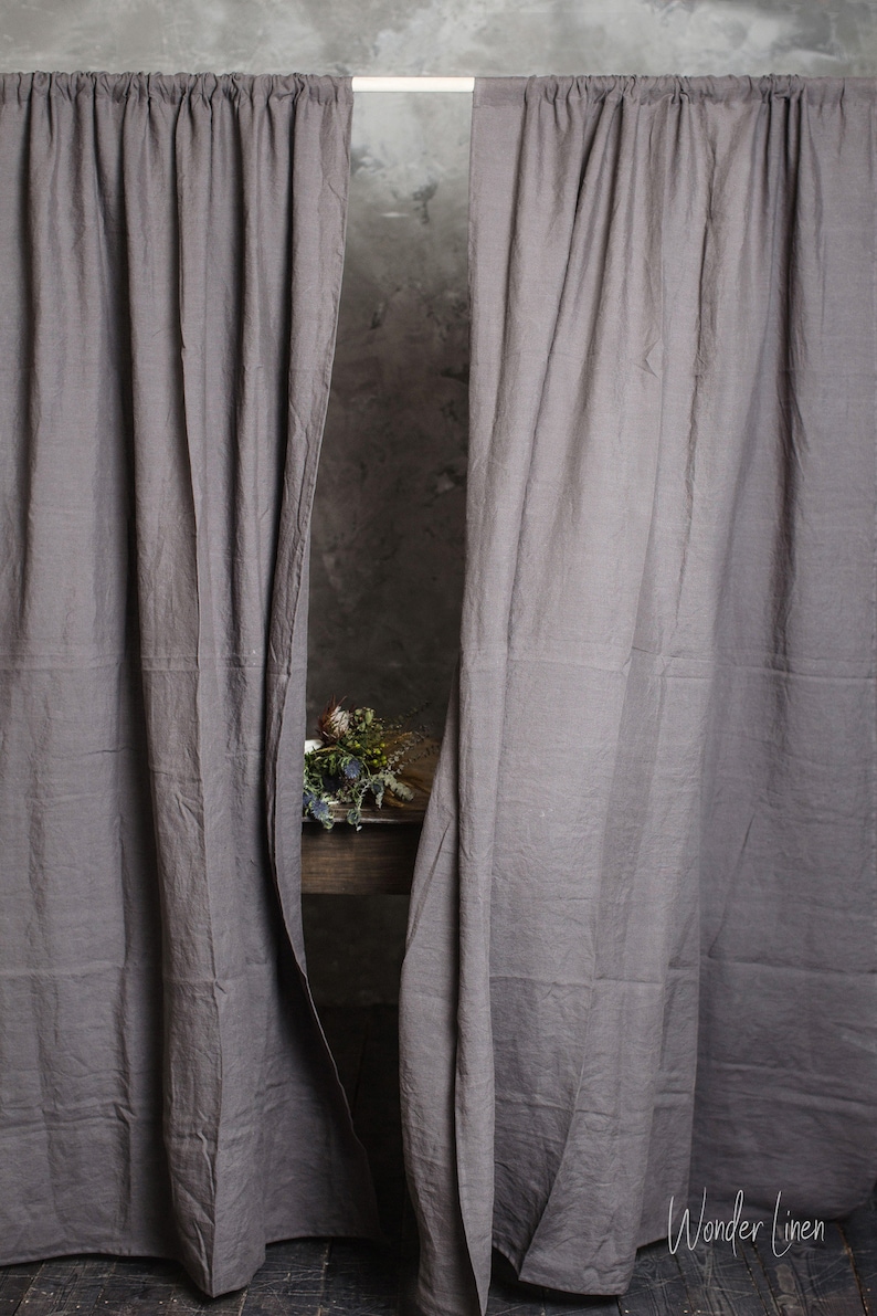 Puede incluir: Dos cortinas de lino gris colgadas de una barra blanca. Las cortinas est&aacute;n ligeramente corridas para revelar una mesa de madera con un ramo de flores secas.
